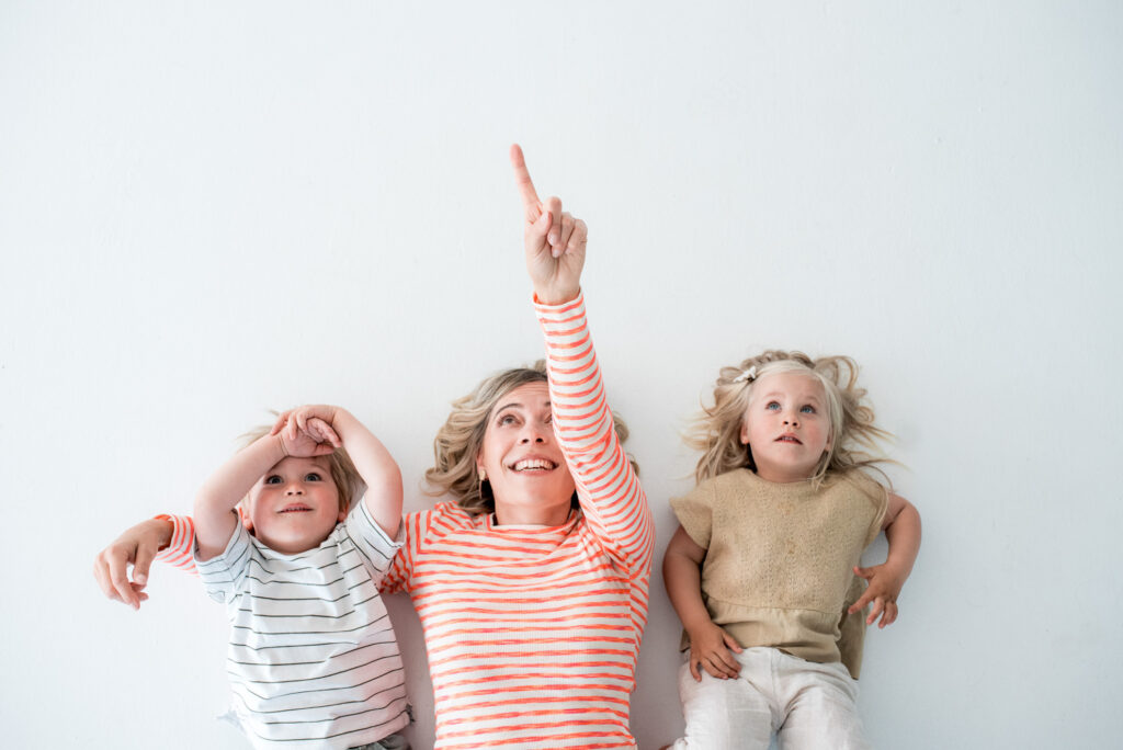 Nina von "Hier kommt Nina!" liegt mit zwei Kindern am Boden und zeigt in die Luft. "Hier kommt Nina" steht für Kindersongs für Groß und Klein.