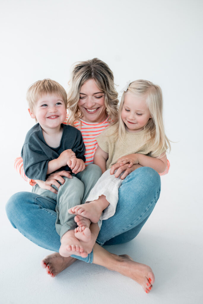 Nina sitzt auf dem Boden und hat zwei Kinder im Arm. Nina singt im Rahmen von "Hier kommt Nina" Kindersongs, die groß und klein alles fühlen lassen.